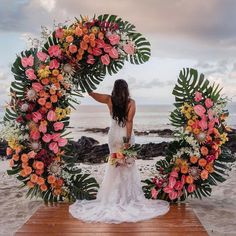 a woman standing in front of an arch made out of flowers and leaves on the beach