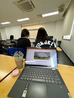 two students sitting at desks with laptop computers on their laps and one student holding a drink in front of them