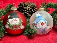 two christmas ornaments with snowmen on them and pine cones in the background, one is red