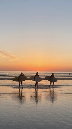 three people holding surfboards while walking on the beach at sunset or sunrise with their backs to the camera