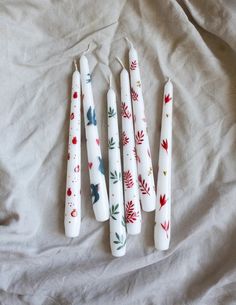 four white candles are lined up on a sheet with red and green leaves painted on them