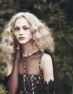 a woman with long blonde hair wearing a sheer top in the woods, posing for a photo