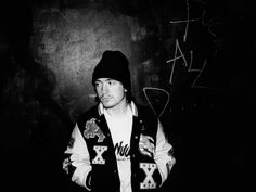 a man standing in front of a black wall with graffiti on it and wearing a beanie