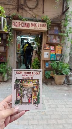 a hand holding up a small book in front of a store with people walking by