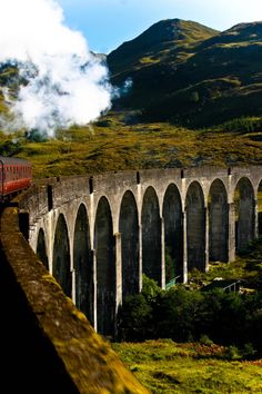 a train traveling over a bridge on top of a lush green hillside next to mountains