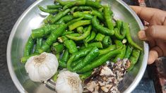 a person holding a metal bowl filled with green peppers and garlic on top of a table