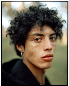 a young man with freckled hair and piercings looks into the camera while standing in a park