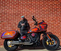 a man standing next to a motorcycle parked in front of a brick wall with an orange and blue design on it