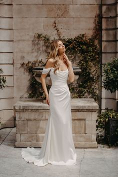 a woman standing in front of a fountain wearing a wedding dress with off the shoulder sleeves