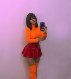 a woman in an orange shirt and red skirt is holding a cell phone while standing against a wall