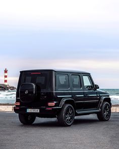 a black mercedes g - class is parked near the ocean with a light house in the background