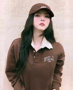 a woman with long black hair wearing a brown shirt and hat standing in front of a wall