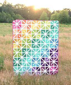 a colorful quilt sitting in the middle of a field with trees in the back ground