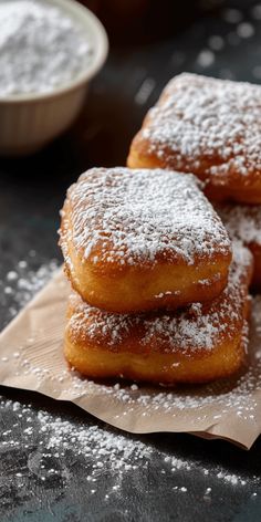 three sugar covered donuts sitting on top of a piece of paper next to a bowl of powdered sugar