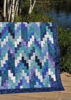 a blue and white quilt sitting on top of a wooden floor next to water with trees in the background