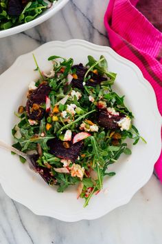 a white plate topped with a salad next to a bowl filled with greens and other vegetables