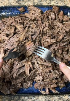 a person is holding a fork and knife over shredded meat on a blue plate, with another hand reaching for it