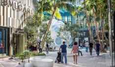 people are walking down the sidewalk in front of shops and palm trees on a sunny day