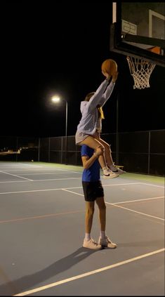 two people are playing basketball on an outdoor court at night, one is holding the ball in the air