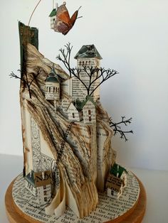 an altered book sculpture with trees and houses on it's sides, sitting on top of a wooden base