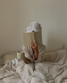 a woman laying in bed reading a book and holding a coffee cup with both hands