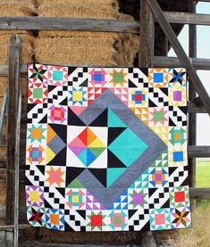 a colorful quilt hanging on the side of a wooden structure with hay bales in the background