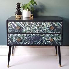 a black and white dresser with palm leaves painted on the drawers, next to a potted plant