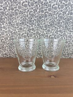 two clear glass cups sitting on top of a wooden table next to a patterned wall