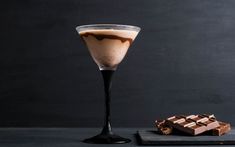 a glass filled with liquid next to pieces of chocolate on a black tablecloth and dark background