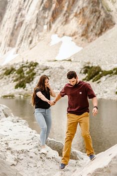 a man and woman holding hands while standing on rocks near water