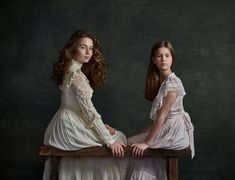 two young women sitting on top of a wooden table next to each other in white dresses