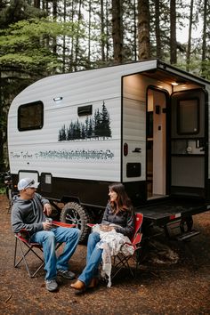 a man and woman sitting in chairs next to an rv parked in the woods near trees