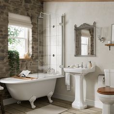 a white bath tub sitting next to a toilet under a bathroom window with a mirror on the wall