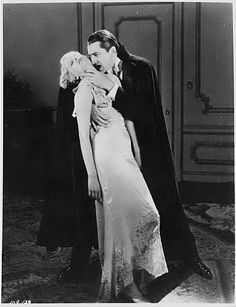 an old black and white photo of a woman in a gown hugging a man wearing a cape