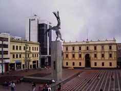 a statue in the middle of a plaza with people walking around and buildings behind it