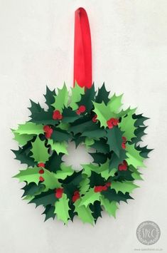 a paper wreath with holly leaves and red berries hanging from a red ribbon on a white wall