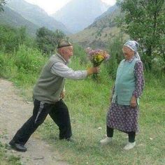 an older couple is standing in the grass with flowers and looking at each other's hand