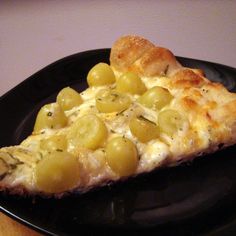 a piece of pizza with cheese and grapes on a black plate next to a wooden table