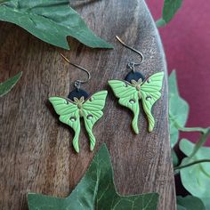 two green and black butterfly shaped earrings sitting on top of a wooden table next to leaves