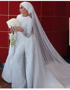 a woman in a white wedding gown and veil with flowers on her head standing next to a red wall