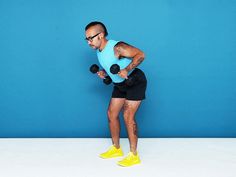 a man in blue shirt and black shorts holding two dumbbells while standing against a blue wall