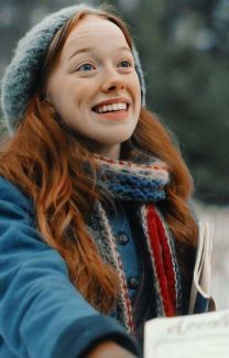 a woman with red hair wearing a gray hat and scarf smiles at the camera while holding a book in her hand