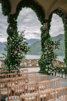 an outdoor ceremony set up with wooden chairs and flowers on the arch over looking water