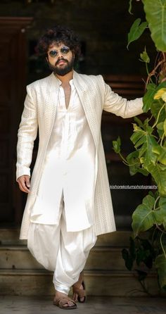 a man in a white suit and sunglasses is standing near some green plants with his hands on his hips