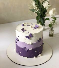 a white and purple cake sitting on top of a table next to a vase with flowers