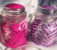 two glass jars with pink and purple leaves painted on the lids are sitting on a white sheet