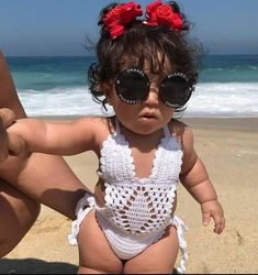 a baby wearing sunglasses on the beach with her mother's arm around her waist