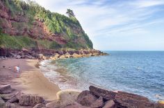 people are walking on the beach next to the water and cliffs in the distance, with one person standing at the edge of the shore