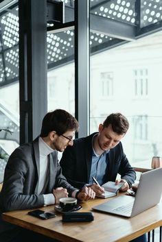 two men are sitting at a table working on their laptops and writing something in front of them