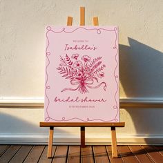 a pink sign with flowers on it sitting in front of a white wall and wooden easel